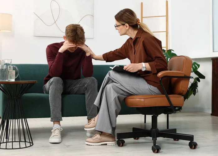 A male patient sits with his head in his hands, appearing distressed, while a female therapist on an office chair rests a hand on his arm to comfort him, both in a modern counseling room.