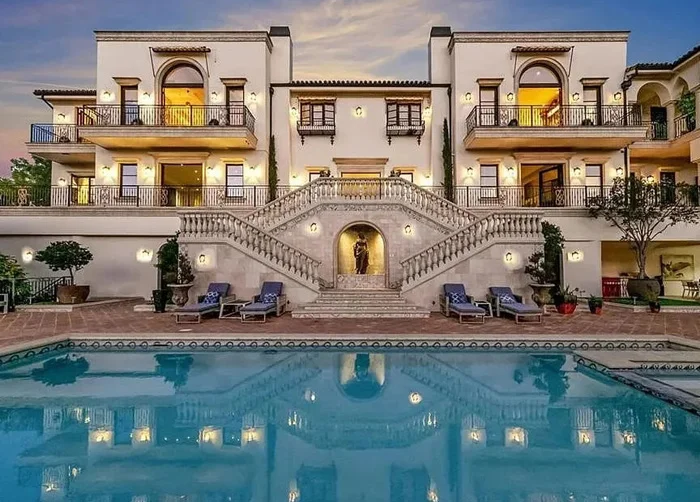 The exterior of a grand luxury estate in Southern California featuring a large swimming pool with perfect reflections, multiple lounge chairs, ornate grand staircases, and a multi-level Mediterranean-style building under a twilight sky.