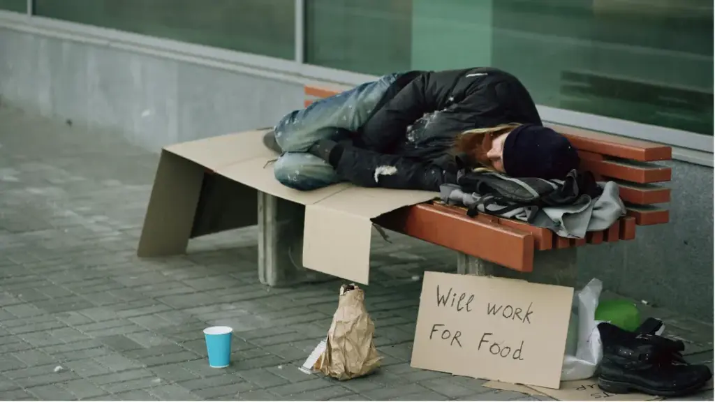 Individual sleeping on a public bench with a sign asking for work, illustrating housing instability linked to chronic methamphetamine use in Los Angeles